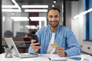 ein Mann ist Sitzung beim ein Schreibtisch mit ein Laptop und ein Zelle Telefon. er ist halten ein Anerkennung Karte und lächelnd foto