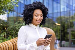 Nahansicht Foto von ein jung Afroamerikaner Frau im ein Weiß Hemd Sitzung auf ein Bank draußen und lächelnd während mit ein Handy, Mobiltelefon Telefon.