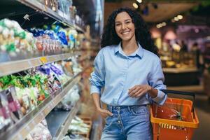 Porträt von glücklich spanisch Frau Käufer im Supermarkt, Frau mit Einkaufen Korb lächelnd und suchen beim Kamera, in der Nähe von Regale mit Produkte im Lebensmittelgeschäft Abteilung foto