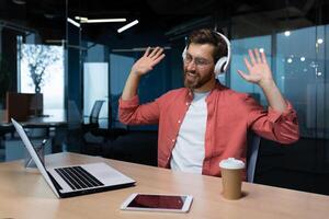 ein Geschäftsmann im ein rot Hemd und Brille hört zu zu Musik- im Kopfhörer, ein Mann Tänze während Sitzung beim das Arbeitsplatz ausruhen, das Boss Verwendet ein Laptop beim arbeiten. foto