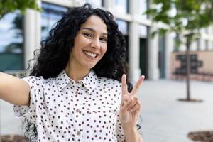 heiter jung Frau mit lockig Haar nehmen ein Selfie, blinkt ein Frieden Zeichen draußen, ausdrücken Freude und Positivität. foto