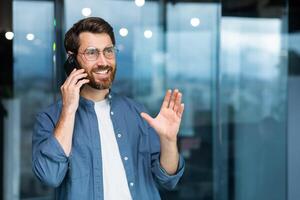 erfolgreich lächelnd Geschäftsmann im beiläufig Hemd reden auf Telefon, männlich Boss im Brille und Bart in der Nähe von Fenster Innerhalb modern Büro. foto