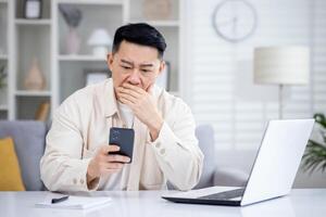 asiatisch reifen Mann Sitzung allein beim Zuhause auf Sofa im Leben Zimmer Kino, Mann empfangen online Benachrichtigung Botschaft mit Schlecht Nachrichten auf Telefon, verärgert Surfen Internet Seiten mit App auf Smartphone. foto