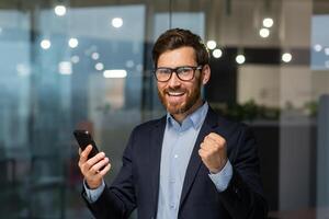 Porträt von erfolgreich Geschäftsmann Investor, reifen Boss im passen und Brille suchen beim Kamera und glücklich feiern Sieg Erfolg, Mann halten Smartphone empfangen online Nachrichten von gut Leistung. foto