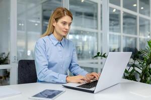 blond Frau im Blau Hemd Sitzung durch Weiß Tabelle und Arbeiten mit Laptop im Licht Arbeitsplatz. besorgt weiblich lesen Geschäft Mail und antworten Fragen von Potenzial International Partner. foto