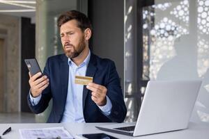 verärgert und enttäuscht jung Geschäftsmann Mann suchen beim Handy, Mobiltelefon Telefon Bildschirm, Sitzung beim Schreibtisch im Büro und halten Anerkennung Karte, Problem mit Konto und Transaktion. foto