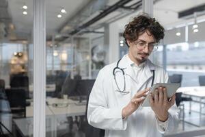 ein Arzt tragen ein Stethoskop und Weiß medizinisch Mantel suchen beim ein Tablette im ein medizinisch Büro. ernst lesen medizinisch Berichte foto