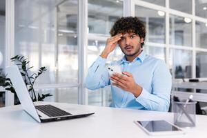 jung spanisch Geschäftsmann Sitzung beim Schreibtisch im Büro, halten Telefon, besorgt halten Kopf und suchen beim Kamera. empfangen Schlecht Nachricht, Mitteilungen, Bedrohungen, Konkurs. foto