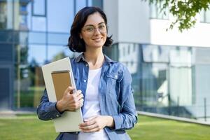 ein jung, heiter Lehrer im Brille und ein Denim Hemd hält ein Notizbuch, Stehen draußen mit ein modern Schule Gebäude im das Hintergrund. foto