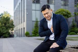 jung asiatisch männlich Geschäftsmann Sitzung gebogen draußen Büro Gebäude im Anzug, halten Hand zu Truhe und Gefühl stark Schmerzen im Herz. foto