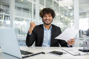 ein heiter jung Mann im ein Geschäft passen feiert ein erfolgreich Projekt oder Leistung während Sitzung beim seine Schreibtisch im ein modern Büro Umfeld, zeigen Sieg Faust und lächelnd mit Unterlagen im Hand. foto