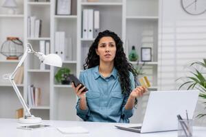 besorgt spanisch Frau im Zuhause Büro halten Anerkennung Karte und Telefon, gegenüber ein Potenzial Betrug Error während arbeiten. foto