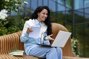 jung schön Frau sitzt auf ein Bank draußen ein Büro Gebäude, Verwendet ein Laptop zu Uhr ein Webinar Kurs , ein Schüler schreibt Daten im ein Notizbuch, Anruf mit ein Lehrer, Entfernung Lernen. foto