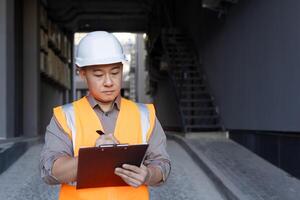 konzentriert und ernst jung asiatisch männlich Architekt, Baumeister. das Ingenieur steht draußen im ein schwer Hut und Weste und macht Anmerkungen im Unterlagen, führt Inspektionen, zeichnet oben ein Projekt planen. foto