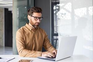 ernst konzentriert Denken Geschäftsmann Arbeiten Innerhalb Büro mit Laptop, reifen erleuchtet Mann im Hemd und Brille beim Arbeitsplatz Tippen auf Klaviatur, Programmierer Entwickler. foto