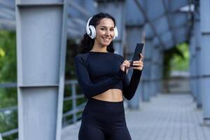 Porträt von glücklich und erfolgreich spanisch Frau, Sportlerin im Kopfhörer und Trainingsanzug suchen beim Kamera, Frau mit online App zum Hören zu Musik, und ausüben draußen. foto