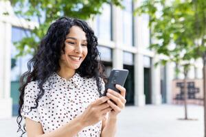 ein jung schön Frau Spaziergänge durch das Stadt mit ein Telefon im ihr Hände, lächelt zufrieden, Verwendet ein Smartphone Anwendung, durchsucht Internet Seiten, Typen ein Text Nachricht, Sozial Netzwerke. foto