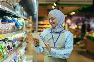 Frau Verkäufer im Super Markt im Hijab mit Tablette Überprüfung Produkte mit Tasche Computer, Muslim Frau in der Nähe von Regale mit Produkte und Waren foto