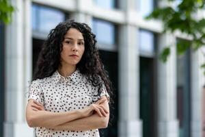 Porträt von ein entschlossen spanisch Frau, Stehen selbstbewusst mit ihr Waffen gekreuzt. das Hintergrund Eigenschaften modern städtisch die Architektur, betonen ein Fachmann Atmosphäre. foto