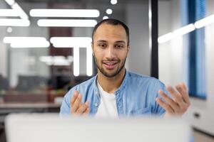 lächelnd jung Mann im beiläufig Büro Kleidung beteiligt im ein online Gespräch, erklären ein Konzept über Forderung. foto