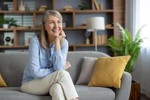 ein heiter Senior Frau sitzt bequem auf ein grau Sofa beim heim, suchen kontemplativ und lächelnd, im ein gut dekoriert Leben Zimmer mit ein modern Stimmung. foto