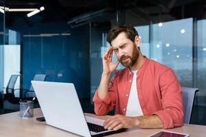 krank Mann beim Arbeitsplatz, reifen Geschäftsmann hat stark Kopfschmerzen, Boss im Hemd funktioniert Innerhalb Büro mit Laptop. foto