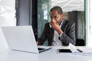 krank Mann beim Arbeitsplatz Innerhalb Büro mit Freude und kalt, Geschäftsmann Niesen Sitzung beim Schreibtisch, afrikanisch amerikanisch Mann mit kalt Verwendet Laptop beim arbeiten. foto