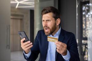 Nahansicht Foto von wütend jung Geschäftsmann im passen Stehen im Büro, halten golden Anerkennung Karte im Hand und suchen enttäuscht beim Handy, Mobiltelefon Telefon Bildschirm.