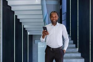 reifen erfahren afrikanisch amerikanisch Mann Innerhalb Büro beim Arbeitsplatz Gehen um Büro mit Telefon im Hand, Porträt von Geschäftsmann im Shirt, lächelnd und suchen beim Kamera mit App auf Smartphone. foto