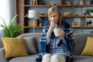 Senior Frau Gefühl nicht wohl während ruhen eingewickelt im kariert Decke und bekommen erwärmt mit heiß trinken im Keramik Tasse. Niesen Dame weht Nase zu Papier Serviette und antiviral Tee drinnen. foto