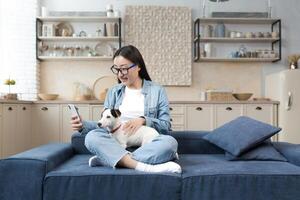 ein glücklich jung schön asiatisch Frau im Brille ist reden auf ein Forderung, halten ein Telefon und ihr Hund im ihr Hände, lächelnd beim das Kamera. Sitzung beim Zuhause auf das Sofa im das Lotus Position. foto