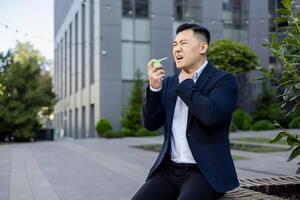 Leiden von ein wund Kehle, ein jung asiatisch männlich Geschäftsmann sitzt auf ein Bank draußen ein Büro Center und Sprays Medizin Aerosol Nieder seine Kehle, halten seine Hand zu seine Nacken. foto