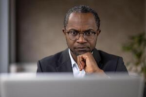ein konzentriert Afroamerikaner Geschäftsmann im Büro Kleidung Arbeiten aufmerksam auf seine Laptop, Anzeigen Professionalität und Einsatz. foto