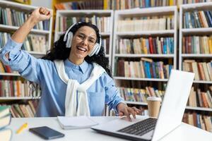 froh jung Frau mit Kopfhörer Jubel im Vorderseite von ein Laptop, erleben Triumph im ein Bibliothek Einstellung. foto
