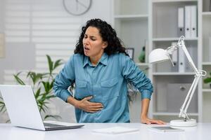 ein mittleren Alters Frau Leiden von stark Bauch Schmerz, Krämpfe oder Beschwerden während Sitzung beim ein Zuhause Büro mit ihr Laptop. foto