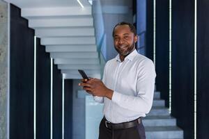 reifen erfahren afrikanisch amerikanisch Mann Innerhalb Büro beim Arbeitsplatz Gehen um Büro mit Telefon im Hand, Porträt von Geschäftsmann im Shirt, lächelnd und suchen beim Kamera mit App auf Smartphone. foto