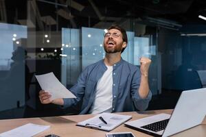 erfolgreich Geschäftsmann im beiläufig Hemd tun Papierkram, Boss mit Bart und Brille Sitzung beim Schreibtisch beim Arbeitsplatz mit Laptop Arbeiten mit Unterlagen, halten Hand oben feiern Sieg und Triumph. foto