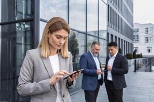 ein erfolgreich und erfahren Geschäft Frau draußen das Büro Gebäude ist lesen von ein Tablette Computer, ein Mitarbeiter mit Kollegen im Geschäft Anzüge auf ein brechen foto