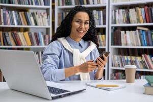 Porträt von ein schön spanisch weiblich Schüler im ein Universität Bibliothek unter Bücher, ein Frau mit lockig Haar ist lächelnd und suchen beim Kamera, halten schärfen, mit ein Anwendung online Lernen. foto