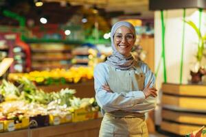 Porträt Frau im ein Hijab wie ein Verkäufer im ein Supermarkt speichern, ein Verkäuferin mit Waffen gekreuzt lächelt und sieht aus beim das Kamera, ein Schürze verkauft Äpfel und Früchte, ein Muslim Frau ist zufrieden im Brille foto
