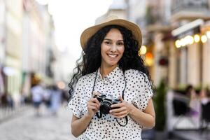 Porträt von jung schön Frau Tourist mit Kamera, spanisch Frau mit lockig Haar im Hut Gehen im Abend Stadt lächelnd und suchen beim Kamera schließen hoch. foto