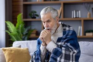 ein Alten kaukasisch Mann mit grau Haar und Bart eingewickelt im ein kariert Decke, Erwärmen seine Hände, Sitzung auf ein Couch im ein modern Leben Zimmer, Gefühl kühl. foto