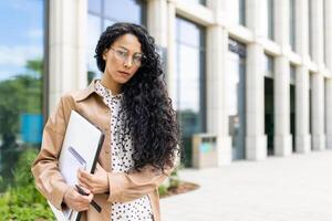 ehrgeizig jung Latein amerikanisch Geschäftsfrau mit lockig Haar halten Unterlagen, Gehen durch Büro Gebäude im städtisch Umfeld. foto