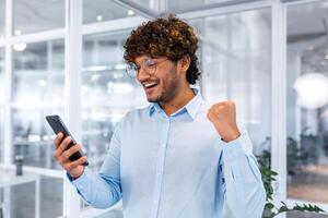 erfolgreich spanisch Programmierer Innerhalb Büro und feiern Sieg und erfolgreich Leistung, Mann lesen empfangen Nachrichten online Telefon, Geschäftsmann im Hemd halten Hand oben Triumph Geste foto