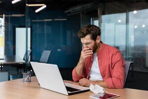 krank Mann beim Arbeit hat Grippe und kalt, Geschäftsmann Niesen und Husten beim Arbeitsplatz Arbeiten Innerhalb Büro beim Schreibtisch mit Laptop beim arbeiten. foto