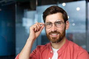 Nahansicht Porträt von jung Geschäftsmann im rot Shirt, reifen Mann im Brille lächelnd und suchen beim Kamera Innerhalb Büro in der Nähe von Fenster, erfolgreich Investor glücklich mit Erfolge und gut Investition. foto