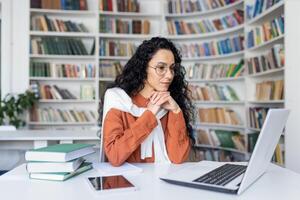 jung ernst weiblich Schüler Denken Über ein Lösung zu ein Abtretung, studieren entfernt während Sitzung im ein Universität Campus Bibliothek, spanisch Frau mit ein Laptop zu bereiten zum Prüfungen. foto