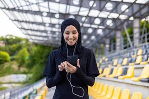 Muslim lächelnd Dame im schwarz Hijab und Sportbekleidung Hören zu Musik- im verdrahtet Kopfhörer während Ausbildung beim Stadion. aktiv Frau mit Telefon Hinzufügen Neu Lied zu Wiedergabeliste zum ausüben auf frisch Luft. foto