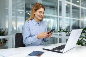 erfolgreich reifen Geschäftsfrau Boss mit Telefon beim Arbeitsplatz Innerhalb modern hell Büro, Büro Arbeiter Tippen online Botschaft auf Smartphone lächelnd foto