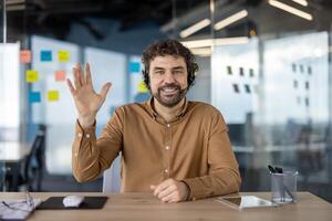 heiter männlich Fachmann mit Headset Gruß im ein zeitgenössisch Büro Raum mit hell, freundlich Haltung. foto